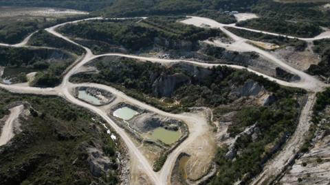An aerial view shows a former china clay pit with pale, dusty tracks winding through excavated land and several small, still pools of greenish water. The site is surrounded by patches of scrub and trees, with exposed rock faces and industrial-looking clearings suggesting past or ongoing quarrying activity.