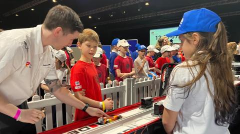There are two children standing either side of a small racing track, getting ready to send their miniature racing cars down. The boy, wearing a red t shirt has the starting trigger in his hand, and a man standing next to him in a white t shirt has his hand on a car, showing him how it works. The other child, a girl in a blue baseball cap and a white t-shirt is watching on, waiting.