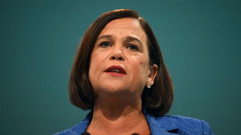Mary Lou McDonald is talking and looking at something above the camera line. She has dark shoulder-length hair and is wearing a blue blazer. The background is solid green. 