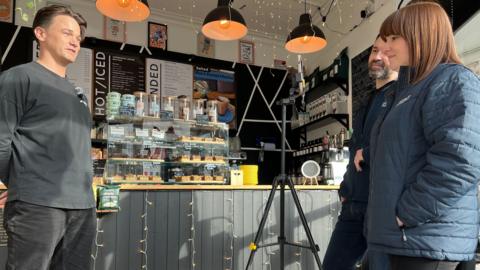 A man stands in a coffee shop talking to a mobile phone held up by a tripod as he is interviewed.