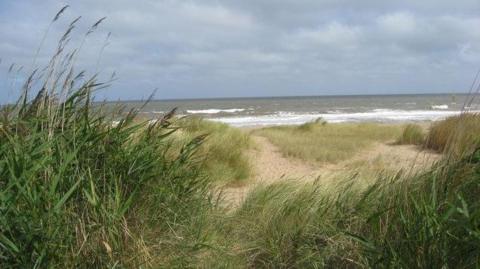 A view of the sea from the sand dunes on a cloudy day, but the sun is shining through. Tall patches of seagrass are blowing in the breeze and the waves are dark with white caps.