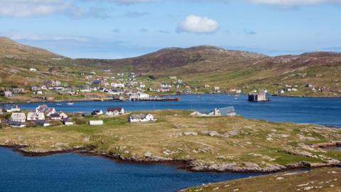 A view across Castlebay on Barra. Houses and other buildings dot a coastal landscape of rocky terrain and blue sea.