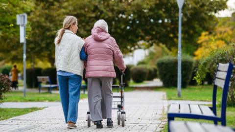 Rear view of senior woman and her home caregiver spending a chilly, windy day outdoors in city park. Autumn walk for elderly patient with walker. - stock photo