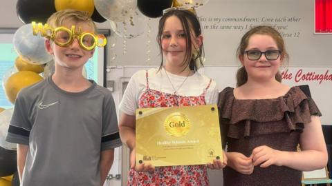Three pupils, smiling and looking at the camera, in a classroom. The girl in the middle is holding a gold certificate, and has on a white top, a necklace and a floral dress. She has long hair tied back, a boy on the left is wearing a grey sports top, a girl on the right is wearing a gold jumpsuit. 