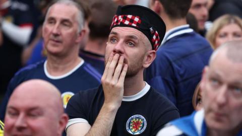 a worried Scotland football fan, wearing a Scotland strip and tartan hat, holds his hand to his mouth