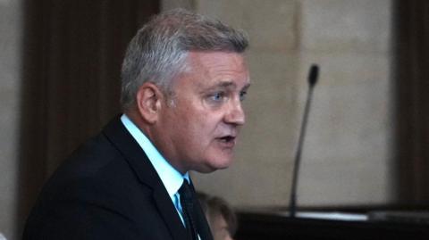 Deputy council leader Russell Quirk, who is wearing a suit and has short grey hair, speaking during a meeting.
