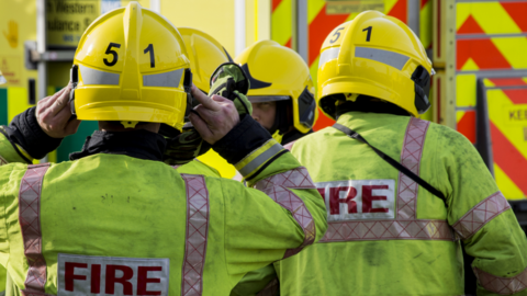 Four firefighters in bright yellow protective equipment and helmets, close up, looking away from camera, with a fire truck in the distance.