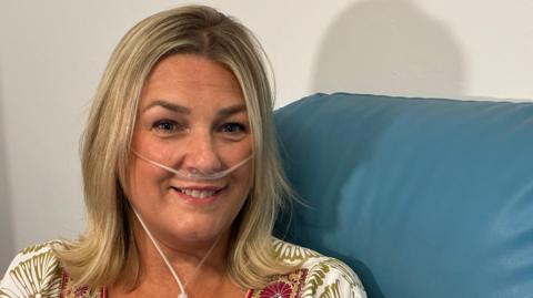 A picture of Helen on a chair in the waiting area of the lung function test area at Nottingham's Queen's Medical Centre. She is smiling at the camera and on display are tubes on her face into her nose which are connected to a portable oxygen machine  