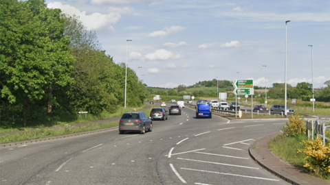 A roundabout on a dual carriageway with multiple vehicles exiting and a signpost pointing the way to Evesham and Stratford and a hospital