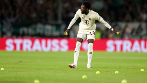 Bayern Munich's Alphonso Davies removes tennis balls thrown onto the pitch by fans