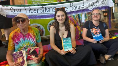 Three women sitting side by side and cross-legged on the floor. Left to right is a woman with sunglasses, a baseball cap and a multi-coloured tie-dye tshirt. She is holding up The Color Purple book and a book by Jeanette Winterson. In the middle sits a smiling woman with long dark hair, a black dress, and sunglasses on her head, holding up a book called Open, Heaven. To her right is a woman with glasses and blonde hair wearing a black top and trousers, holding a book called Women