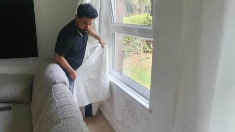 Man pulling back curtain with damp wall underneath a window