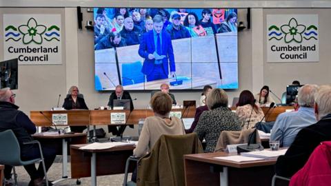 Dozens of councillors are gathered for a meeting in a white room with wooden desks and signs on the wall reading: "Cumberland Council". A large screen behind the head table shows Josh Kirkwood standing and reading from a piece of paper. There are dozens of people sitting in the public gallery behind him.