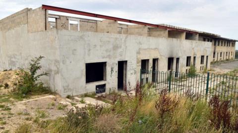 A large two storey building which resembles a building site. The building has no roof and exposed steel girders on the second floor. Windows have no glazing and doors are empty.
