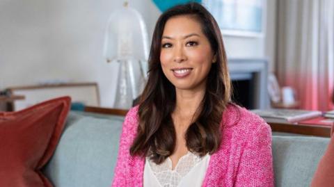 Vivien Wong wearing a white lace camisole and bright pink wool cardigan sitting on a grey sofa in a plush home