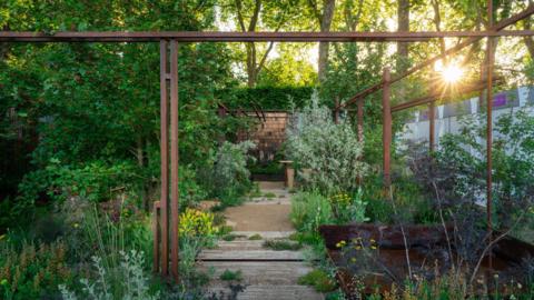 The image shows a garden bathed in sunlight. A narrow gravel-and-stone pathway leads through dense greenery toward a small wooden structure in the background. Tall rust-colored metal frames form an open pergola-like structure overhead, creating strong geometric lines that contrast with the organic plants.