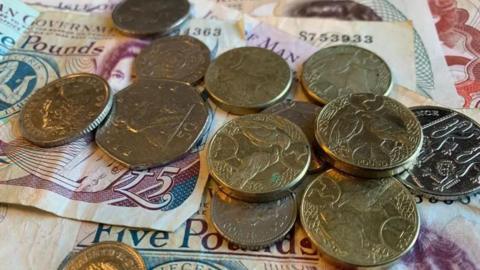 A collection of coins on top of Manx banknotes. There are gold £1 coins, and silver 50p, 20p, and 10p coins lying on top of £5 and £10 notes.