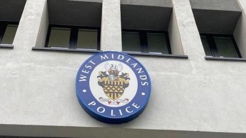 A grey concrete building with gaps for windows with dark frames. On the front is a blue circular badge with the words "West Midlands Police" around the edge and a crest in the middle