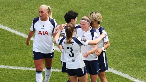 Four Tottenham players, wearing white shirts and dark shorts, gather around Signe Gaupset, with her mouth open and a white headband on her blonde hair, to celebrate her goal