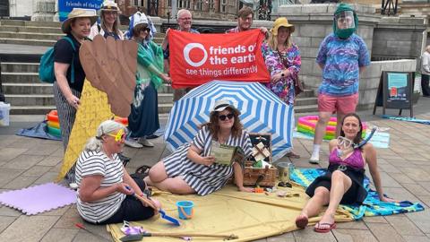 Campaigners dressed in bathing costumes and carrying a red Friends of the Earth banner sit and stand around a yellow bathing towel