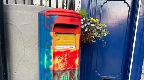 The photograph shows a Guernsey postbox that had been painted in the colours of the rainbow. There is now blue spray paint up the left side of it and red spray paint up the right side of it and on the front.  