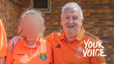 A man in a bright orange top smiles at the camera. He has his arm round a woman whose face has been blurred out. They are standing in front of a brick wall.
