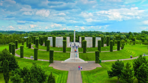 A memorial in the middle of wooded countryside. A white monument can be seen in the distance with white walls on either side and white steps leading up to it. Footpaths encircle the memorial along with a number of vertical hedges.
