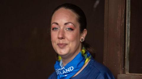 Sheree Gregg smiles at the camera. She has her dark brown hair tied back and wears pink eyeshadow. She has a blue and yellow Team Scotland scarf and wears a blue -tshirt.