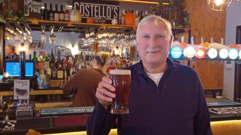 A man with short white hair and a white moustache, wearing a blue jumper, stands in front of a bar while holding up a pint in his right hand.