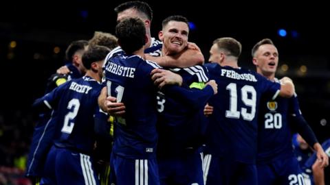 A group of footballers in navy jerserys cuddle and celebrate following a goal. 