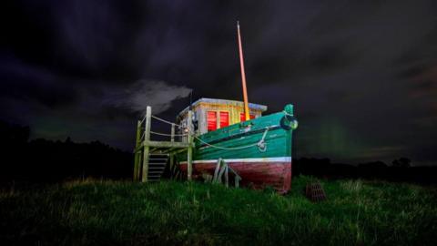 An old, weathered fishing boat, painted green and red, resting on land amidst short, green grass. A small wooden structure resembling a cabin has been built onto the boat's deck. The cabin's windows glow with a warm, reddish light. The boat sits amongst a dark sky