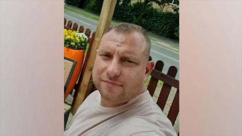A man with short brown hair who is muscular has a pale pink top on and is looking into the camera with a serious expression. Behind him is a wooden fence and then a road behind that. He appears to be sat down at a table.