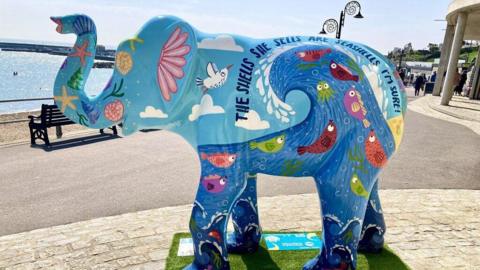 A fibreglass elephant stands on a platform next to a beach. It appears to be about twice the height of a nearby bench. It is painted mainly in blue, with fish and sea creatures.