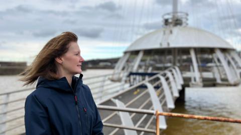 Deanne Shallcross, CEO of Whitehaven Harbour Commissioners. She is stood at Whitehaven Harbour by the walkway that leads to the Crow's Nest - a pagoda-type white structure on the water. She is looking out at the harbour, side-on with the wind blowing back her shoulder-length brown hair. She is wearing a blue waterproof jacket. The background is blurred.
