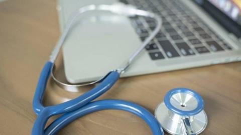 A laptop on a wooden desk with a blue stethoscope draped over it 