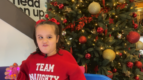 Cleo playing with a toy. She is wearing a red jumper that reads Team Naughty and standing in front of a Christmas tree. 