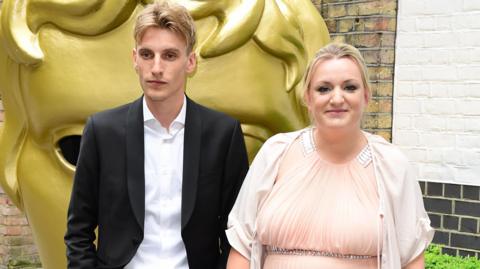 Charlie and Daisy May Cooper. They are both blonde and at the BAFTA awards. Charlie is wearing a white shirt and a black suit jacket. Daisy May is wearing a pink dress with silver sparkly details. Daisy May is looking at the camera and smiling. Charlie is looking slightly off to the side.