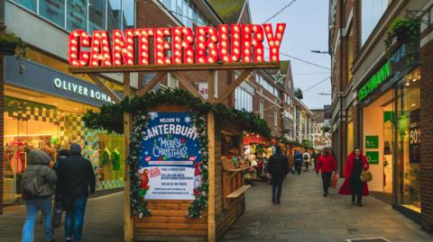 A stall decorated with a large illuminated sign saying "Canterbury" on top of a wooden hut in a city street decked out for Christmas, as shoppers walk past.