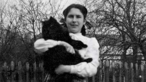 A black and white photograph of a dark-haired girl wearing a white blouse and holding a large black cat.