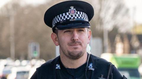 Paul Hogben wearing a black police hat and the black uniform with a walkie-talkie attached to his chest. He is walking by a road with queuing traffic.