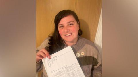 A smiling woman stands holding a GCSE certificate.