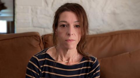 Emma Simmonds is sitting down on a brown sofa. She is looking directly at the camera and is wearing a navy and brown striped long-sleeve T-shirt. She has brown hair, pulled back in a ponytail.