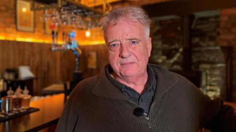 A man with short grey hair, wearing a navy shirt and and a dark green, half zip jumper. He is standing in front of a bar.