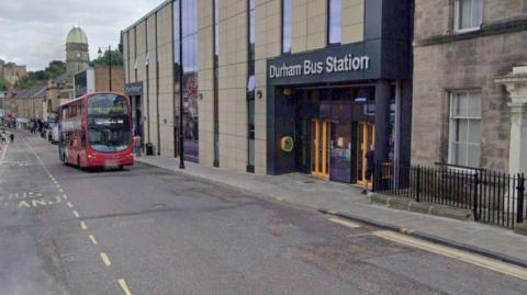 A Google Streetview screenshot of North Road outside the bus station in Durham. A double decker bus is travelling down the road on the right-hand side. The left side of the road has road markings for a bus stand all the way along it. 