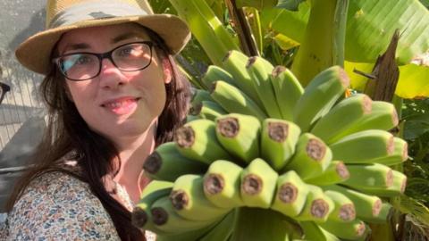 A woman with glasses and a hat posing next to a bunch of bananas