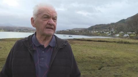 Drew Miller is an older man with white hair. He is standing outdoors near a loch, hills and a scattering of white houses. He is wearing a dark jacket over a dark blue polo shirt.