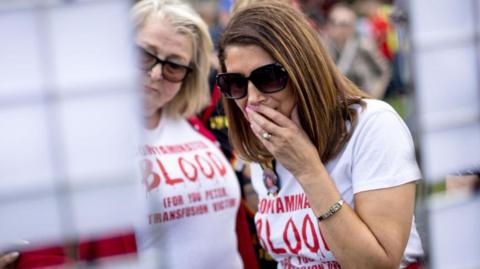 Two women stand outside the building in Westminster where the final report into the infected blood inquiry was published in May 2020. They are both wearing white T-shirts with the slogan 'Contaminated Blood" written in red ink. The women in the right is wearing dark sunglasses and holding her face to her mouth. 