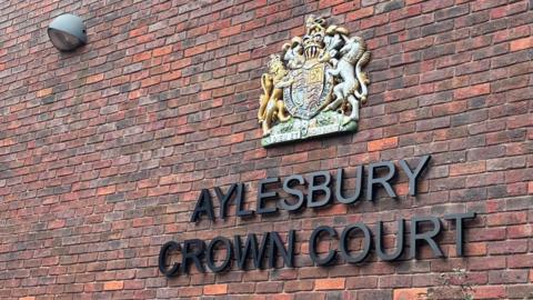 A brick wall belonging to Aylesbury Crown Court. The building is red bricks and there is lettering on the wall and a crest. The lettering is black and says AYLESBURY CROWN COURT.