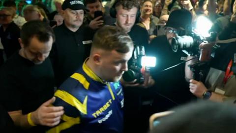 A man in a blue and yellow top walking through a crowded bar with camera lights on him