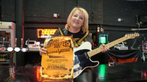 Mayor of Wolverhampton Councillor Linda Leach pictured smiling and holding a black and white electric guitar. She has short blonde hair and is holding an orange poster in her left hand that says Mayors Fest 2025.